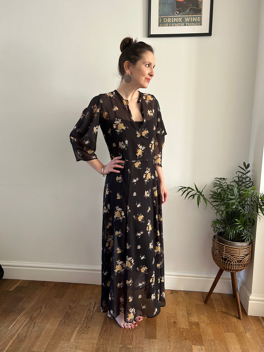 Woman wearing a floaty maxi dress featuring cowl sleeves in a black chiffon fabric with yellow flowers.