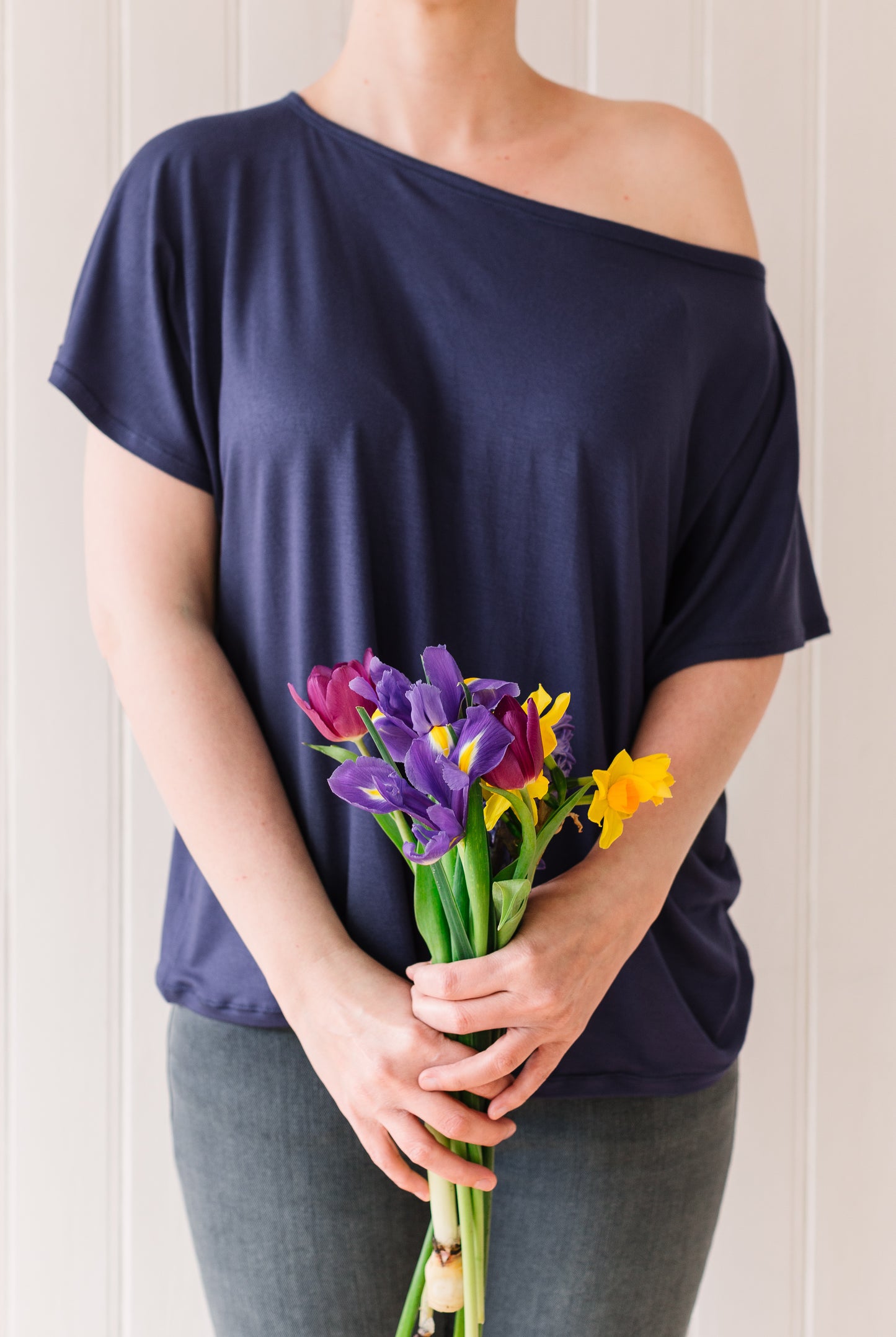 Front view of a woman wearing an off the shoulder slouchy t-shirt in a midnight blue fabric holding a bunch of flowers.